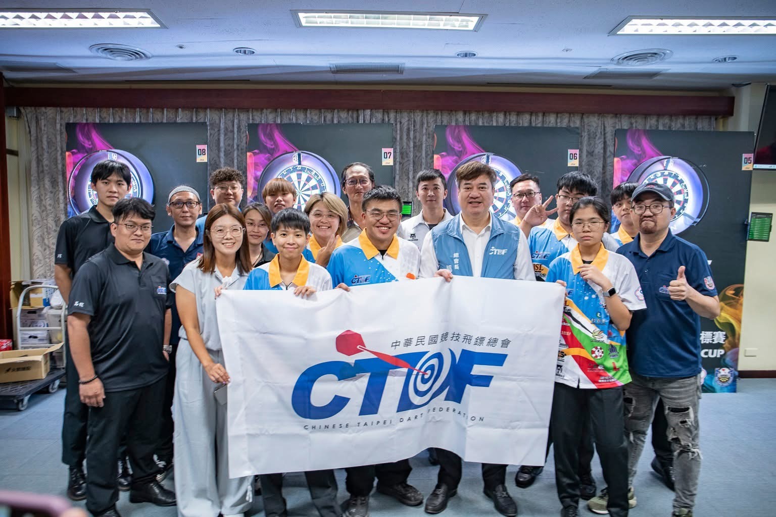 中華民國競技飛鏢總會(CTDF)代表與參賽選手、工作人員於競技飛鏢賽事現場拉起會旗的歡樂大合照，展現推廣飛鏢運動的熱血與凝聚力。