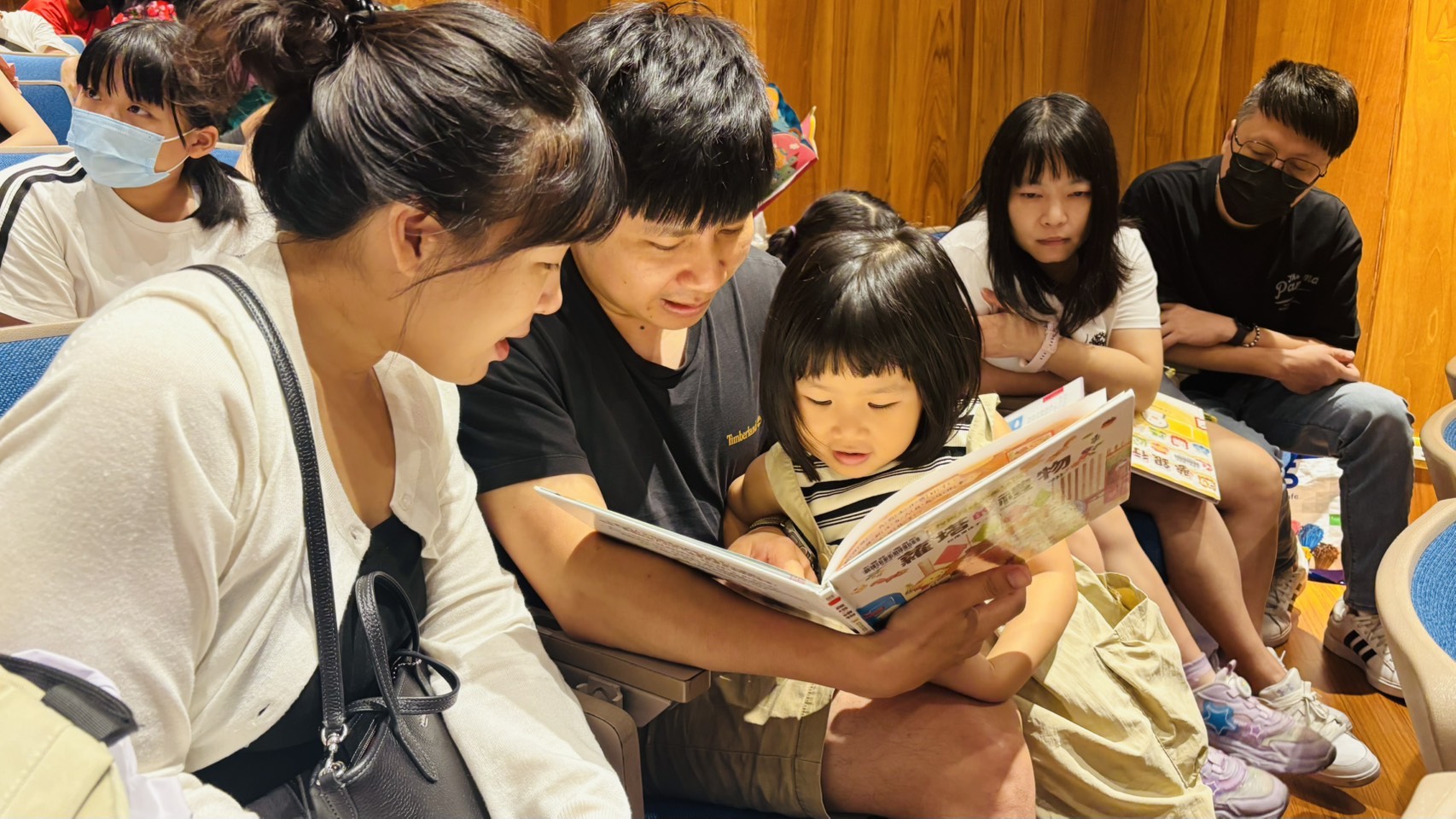 父母將年幼孩子抱在懷中共同翻閱圖書,孩子專注看著書中內容,呈現親子閱讀融入家庭日常的溫暖畫面。